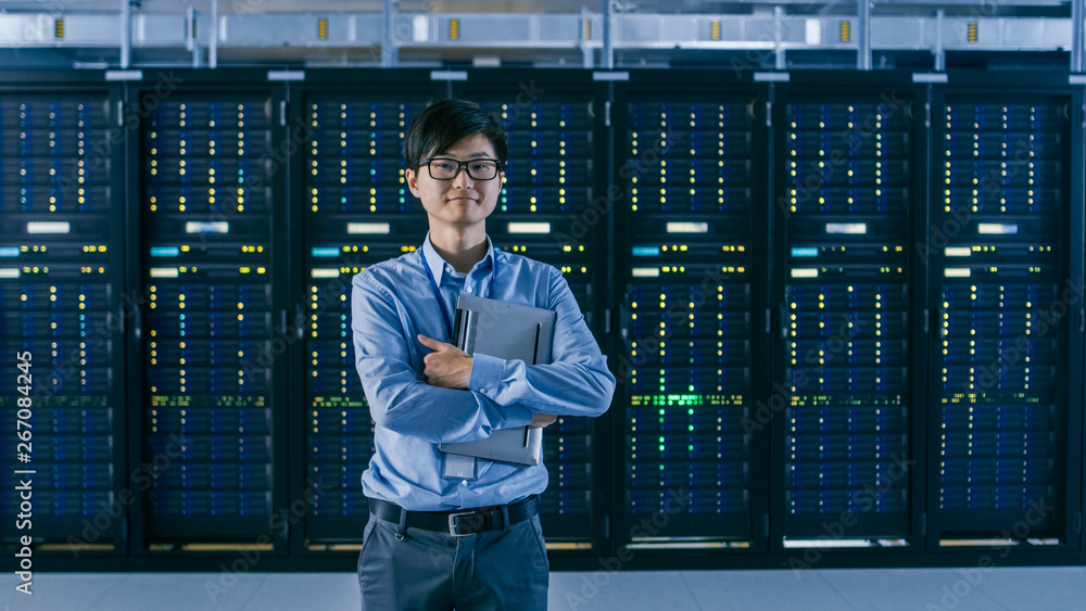  In the Modern Data Center: Portrait of IT Engineer Standing with Server Racks Behind Him, Holding L
