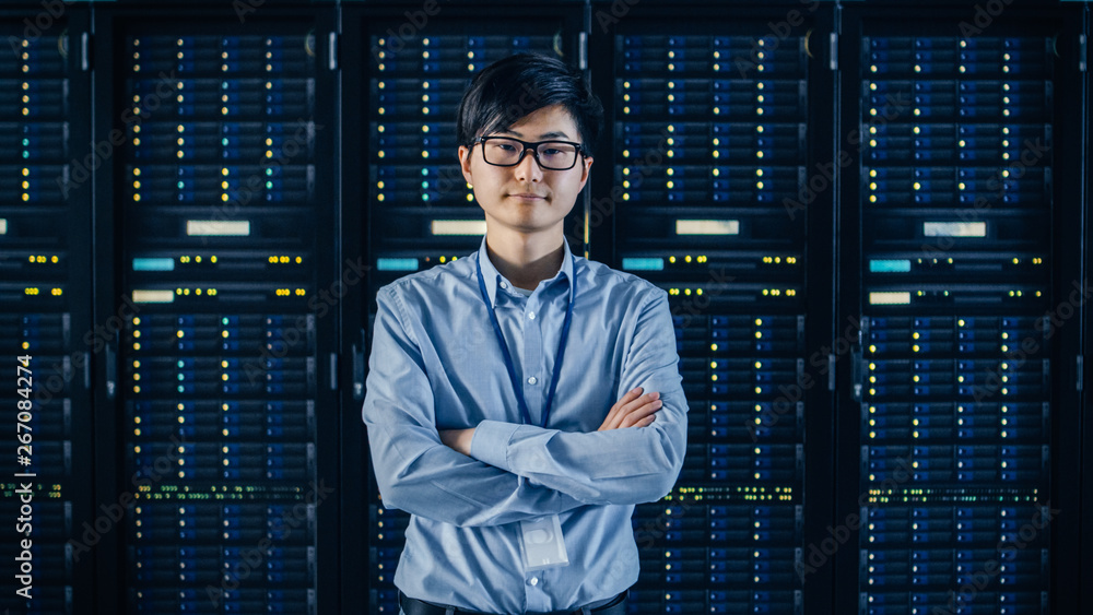  In the Modern Data Center: Portrait of IT Engineer Standing with Server Racks Behind Him, Crossing 