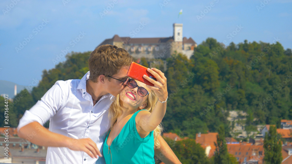 CLOSE UP: Happy man kisses his girlfriend on the cheek while taking selfies.