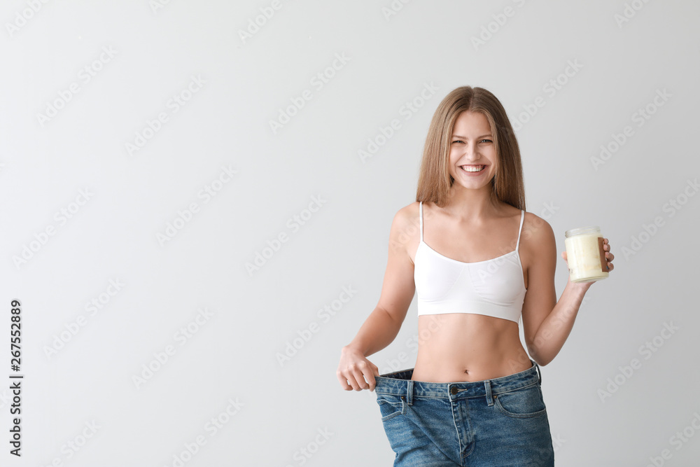 Beautiful young woman in loose jeans and with jar of coconut oil on light background. Weight loss co