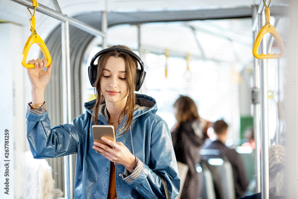 年轻女性乘客在现代有轨电车中移动时戴着耳机和智能手机站着