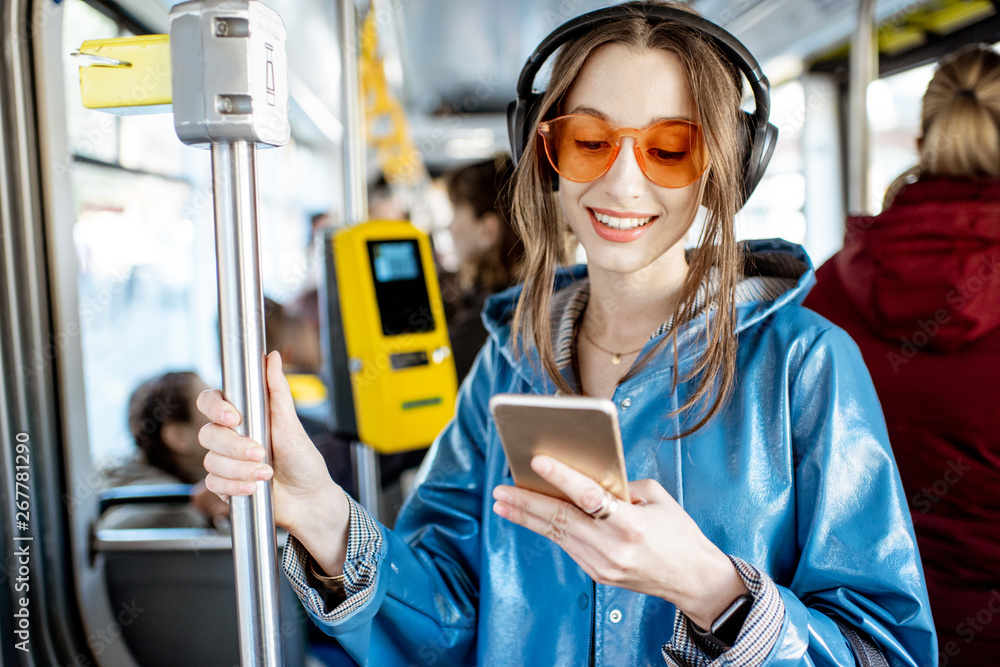 Young stylish woman using public transport, standing with headphones and smartphone while moving in 