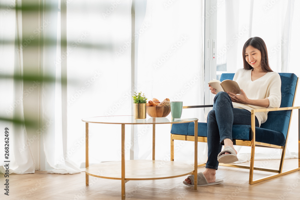 Young beautiful Asian woman relaxing in living room at home, reading books, drinking coffee in the m