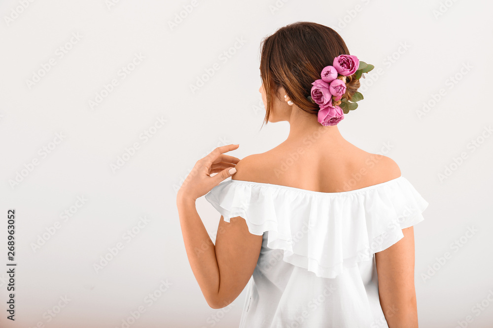 Young woman with beautiful flowers in her hair on light background, back view