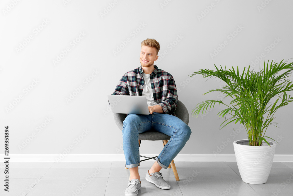 Handsome young man with laptop sitting on chair near light wall