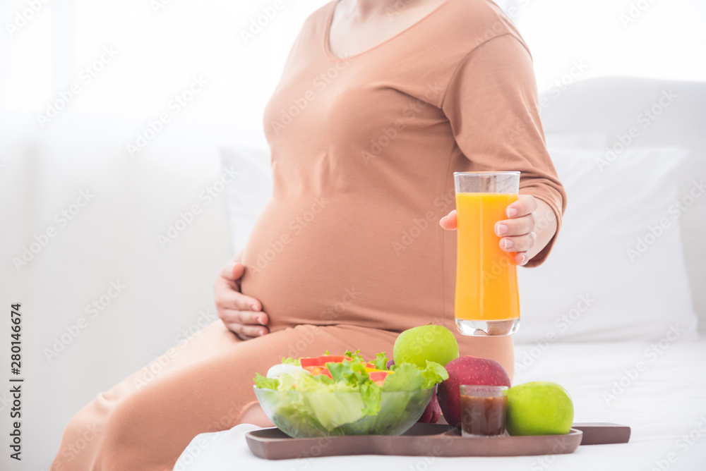 孕妇坐在床上，手里拿着一杯橙汁。把注意力集中在橙汁杯上。
