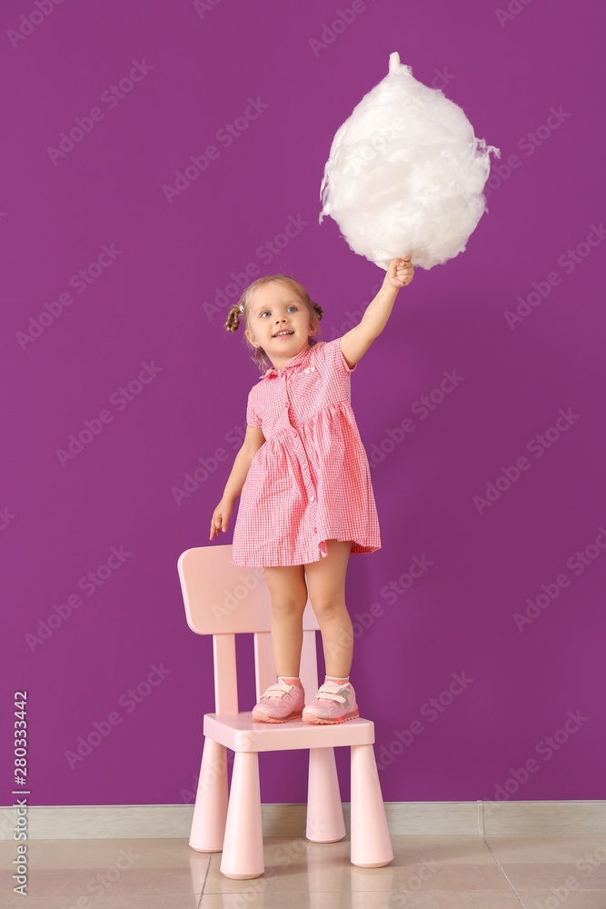 Cute little girl with cotton candy standing on chair against color wall