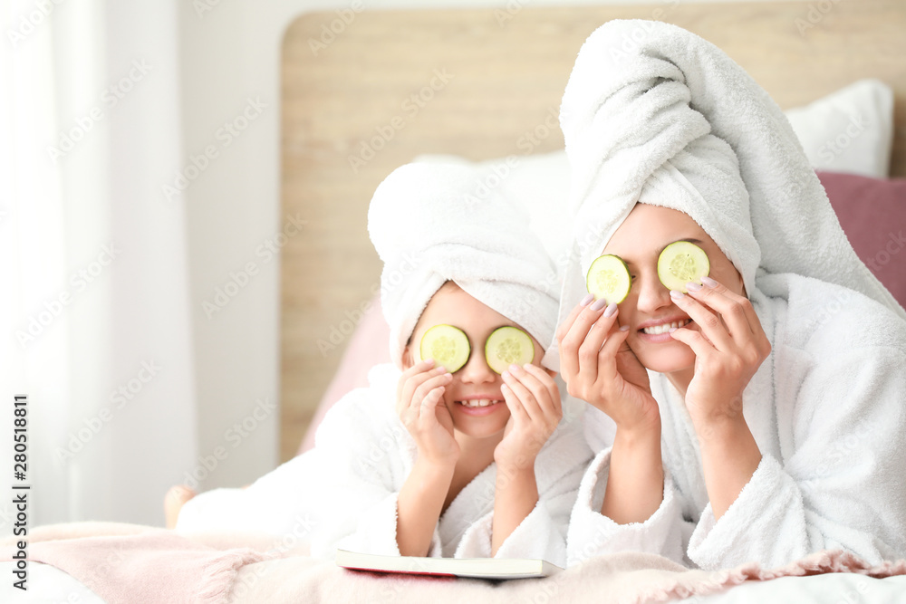 Mother and her little daughter in bathrobes and with cucumber slices in bedroom