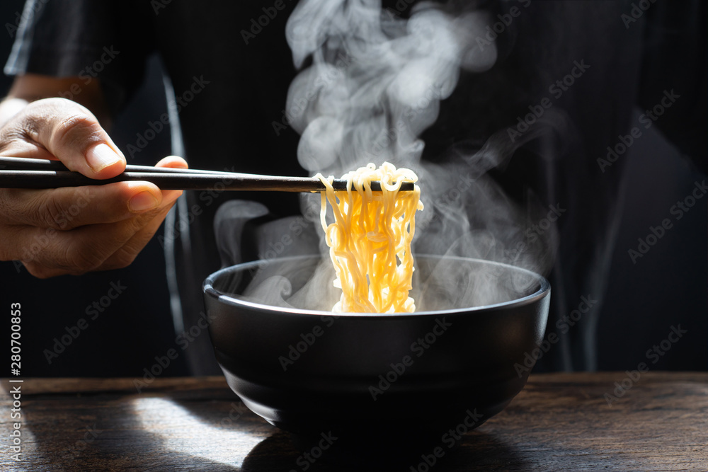 Hand uses chopsticks to pickup tasty noodles with steam and smoke in bowl on wooden background, sele