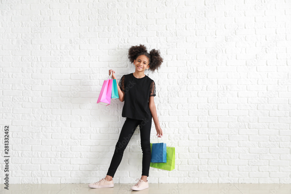 时尚的非裔美国女孩，穿着牛仔裤，在白砖墙附近拿着购物袋