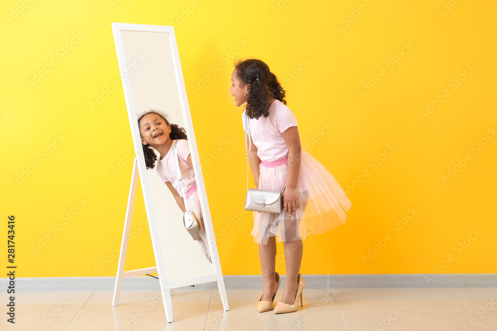 可爱的非洲裔小女孩，穿着妈妈的鞋子，看着彩色墙附近的镜子