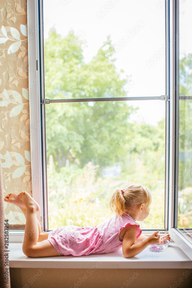 漂亮的金发女孩穿着粉色连衣裙，躺在家里窗台上敞开的大窗户附近玩耍