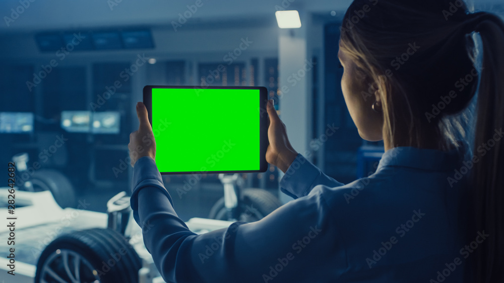 女工程师拿着一台带绿屏实物模型的平板电脑，看着电动车底盘