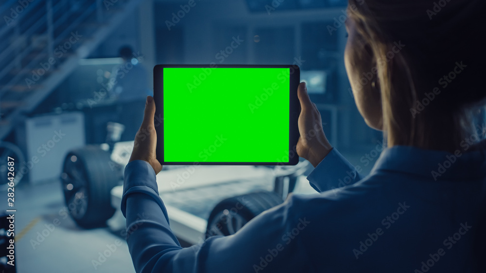 Female Engineer Holds a Tablet Computer with Green Screen Mock Up and Looks at Electric Car Chassis 