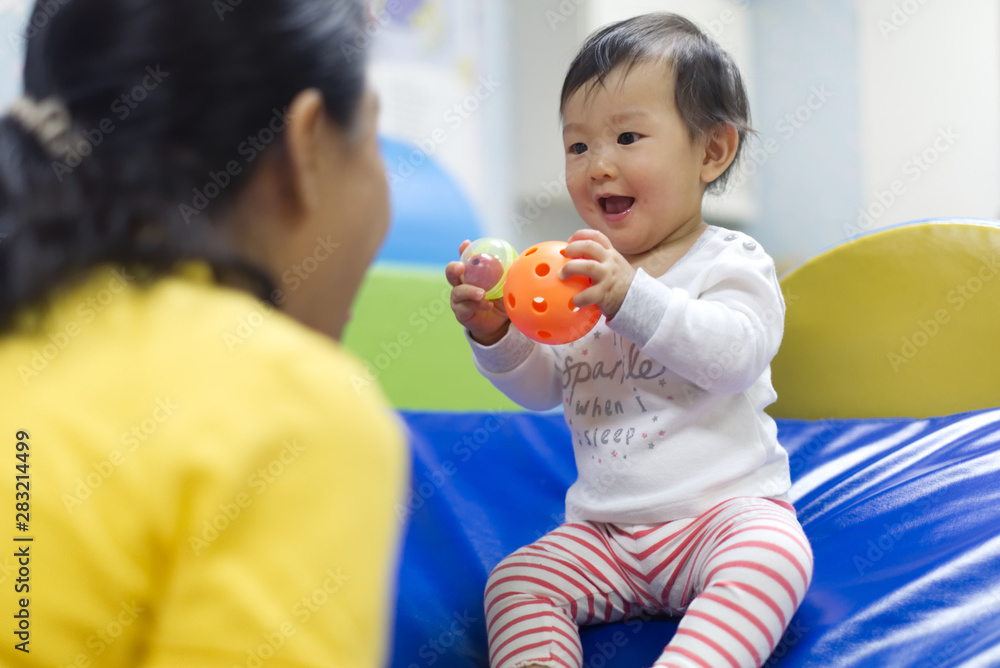 Young little Asian baby holding balls and toys playing with female preschool teacher in classroom. K