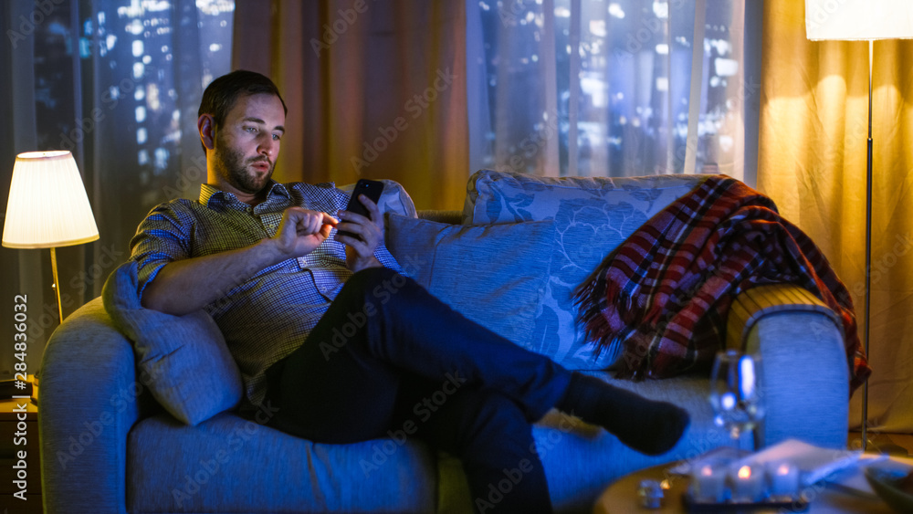 晚上，中年男子坐在沙发上使用智能手机。打开电视照亮他。在