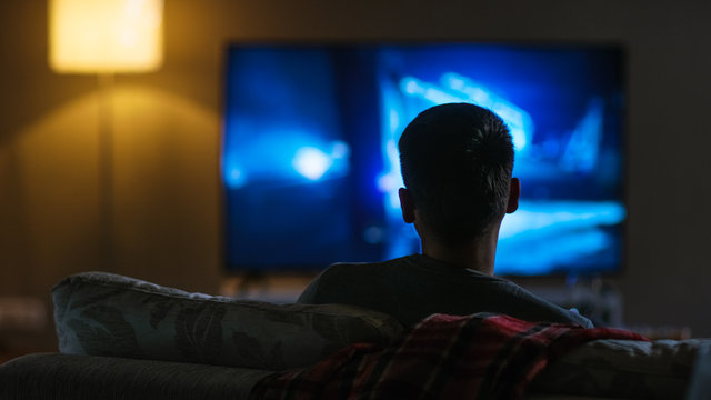 一个男人坐在沙发上用他的大平板电视看电影的后视图。
