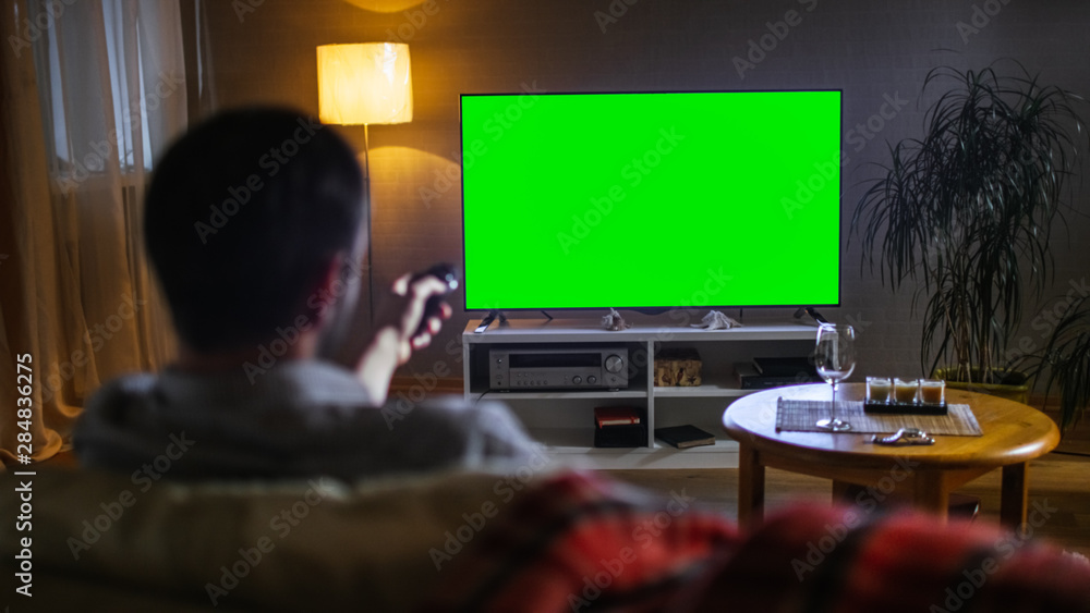 在一个中年男人坐在沙发上看大平面电视的傍晚背影中，他挥了挥