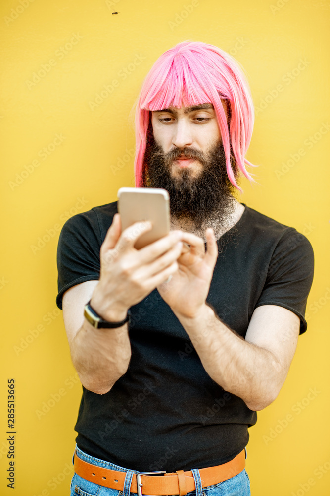 Funny portrait of a stylish man with beard and long color hair usning smart phone on the yellow back