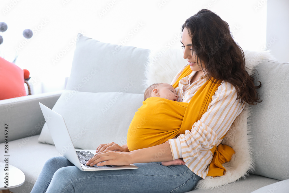 Young mother with little baby in sling using laptop at home