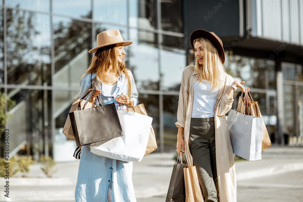 两个快乐的女朋友拿着购物袋走在商场前，感觉很满足。