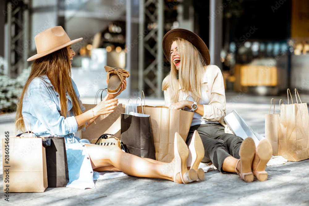 两个快乐的女朋友坐在购物中心附近，手里拿着购物袋和新买的东西