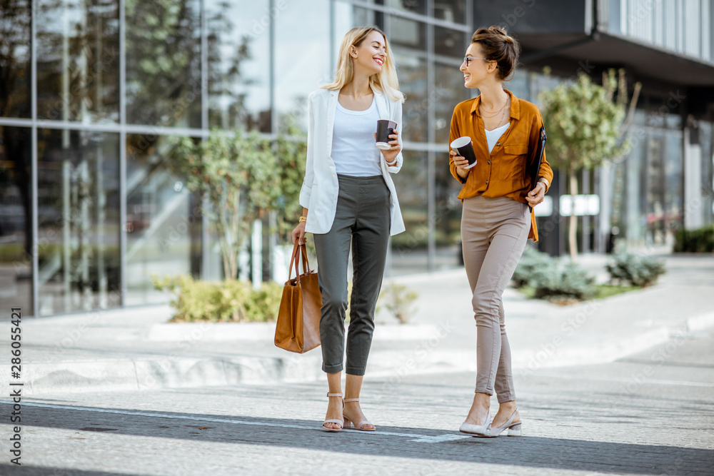 Full body portrait of a two young businesswomen walking with coffee cups near the modern office buil