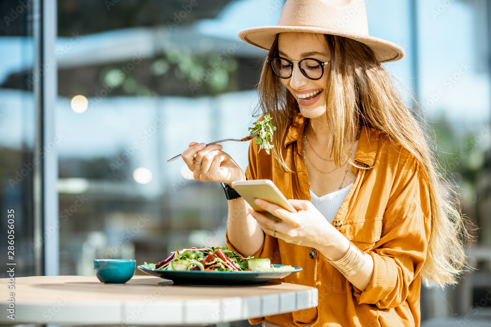 时尚的年轻女子在餐厅露台上吃着健康的沙拉，在夏天感到幸福