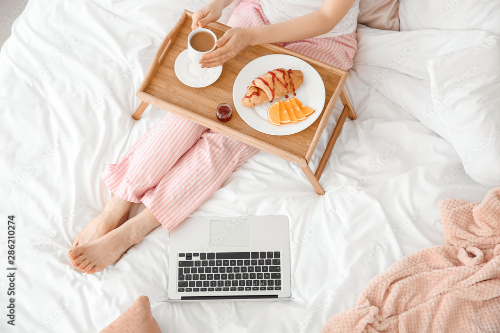 漂亮的年轻女人拿着笔记本电脑在床上吃早餐