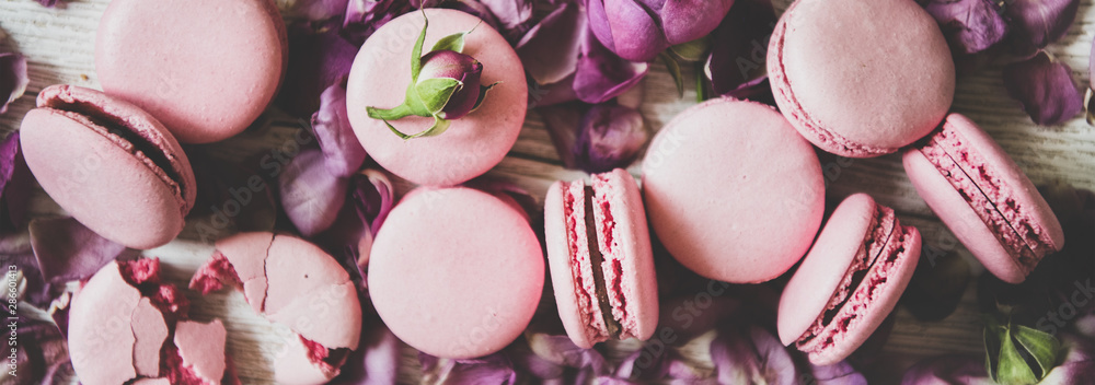 Flat-lay of sweet pink macaron cookies and rose buds and petals over wooden background, top view, cl