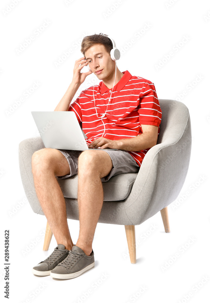 Young man with laptop listening to music while sitting in armchair against white background