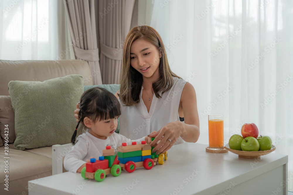 Asian toddler girl with mother play with colorful wooden block train toys on table at home.