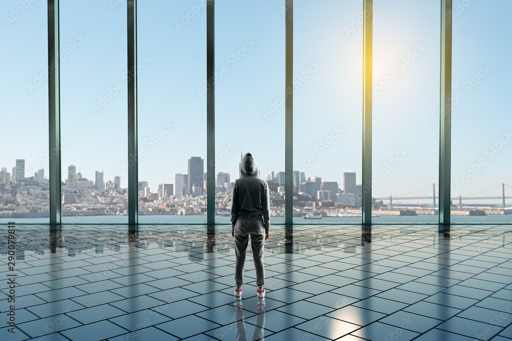 在空荡荡的宽敞大厅里，一位穿着运动服的女士，有玻璃墙，可以看到大都市的美景。