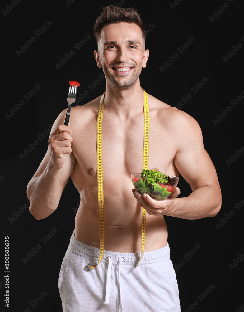 Handsome muscular man with measuring tape and salad on dark background. Weight loss concept