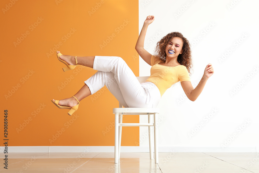 Fashionable young African-American woman sitting on chair against color wall