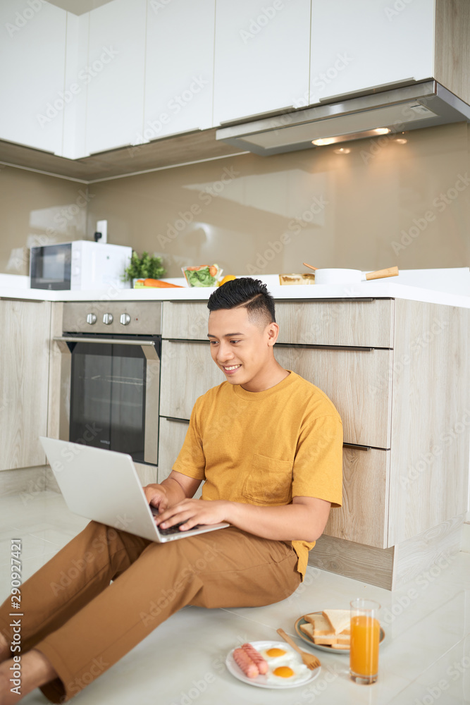 专心的亚洲男子坐在厨房地板上使用笔记本电脑