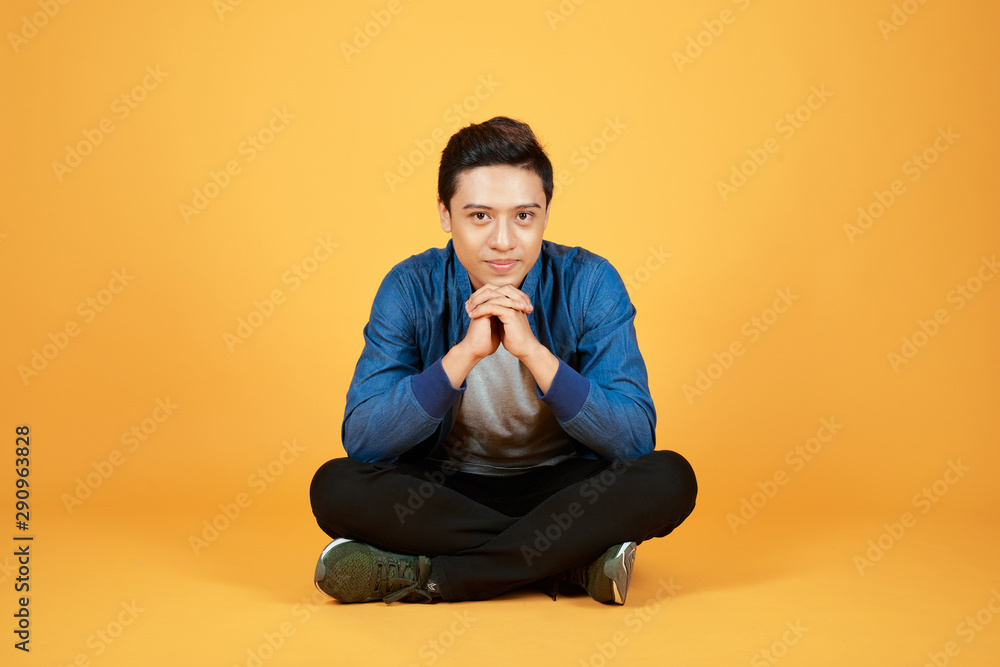 长相英俊的年轻亚洲男子盘腿坐在地板上，被隔离在橙色背景上。