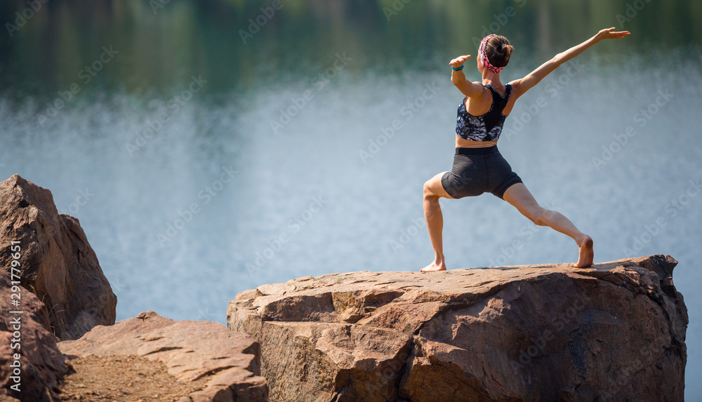 年轻女子在湖景优美的岩石上的晨间公园里做瑜伽。瑜伽女孩