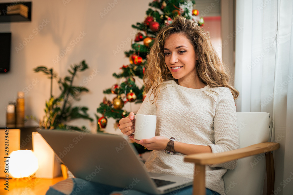 在家享受圣诞节。一个年轻女人在家看着笔记本电脑喝茶的画像。