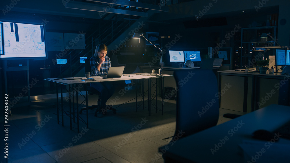 美丽的女工程师坐在办公桌前在笔记本电脑上工作。蓝图躺在桌子上