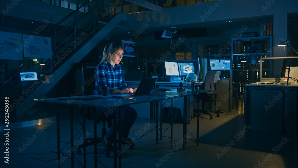 美丽的女工程师坐在办公桌前用笔记本电脑工作。蓝图躺在桌子上