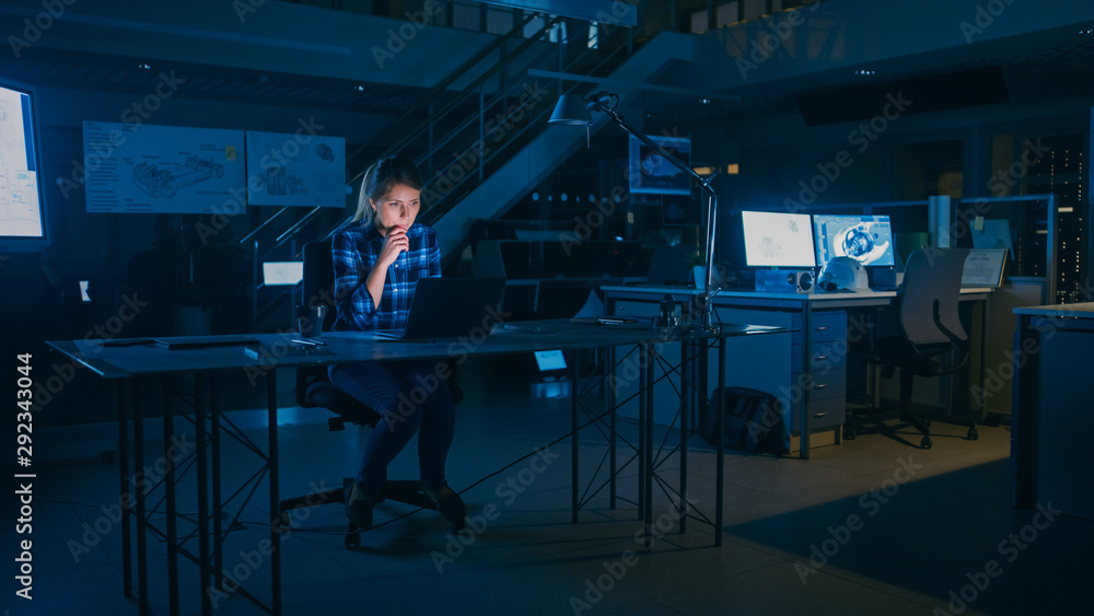 美丽的女工程师坐在办公桌前用笔记本电脑工作。蓝图躺在桌子上