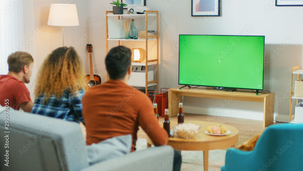 三个朋友坐在家里的沙发上，一边吃零食一边看绿色Chroma键屏电视