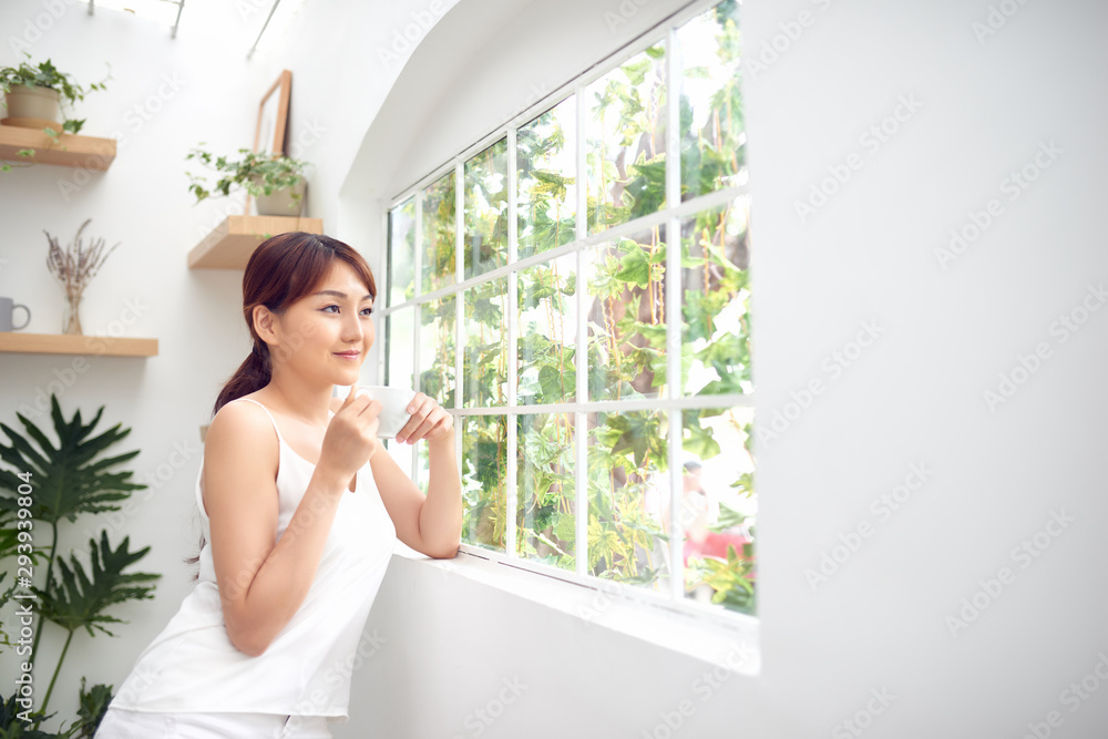 年轻漂亮的女人站在窗户后面喝咖啡，看着外面享受休息
