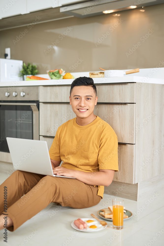 年轻的商人面带微笑，在明亮的厨房里吃早餐，用笔记本电脑工作。