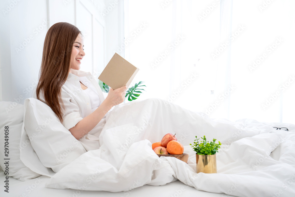 美丽的亚洲女性白色连衣裙长发在白色床上看书，有蛤和欢快的白色卧室