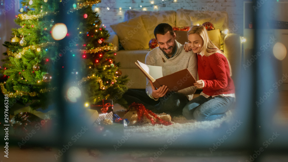 透过窗户看。美丽可爱的情侣坐在圣诞三号树下，手里拿着礼物和Rea