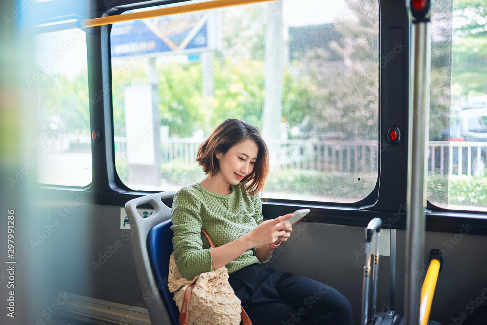 美丽快乐的年轻女子坐在城市公交车上，看着手机