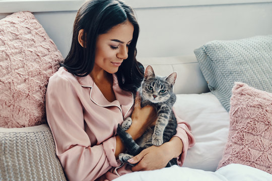 美丽的年轻女子穿着睡衣微笑着拥抱家猫，而在家里的床上休息