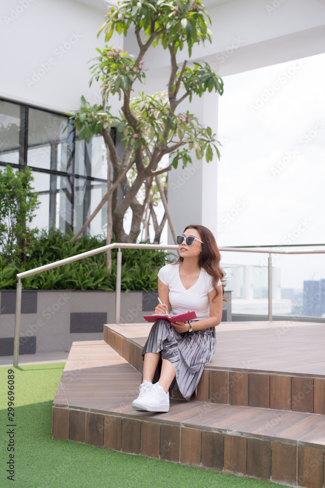 Happy young woman wearing sunglasses sitting on stairs and writing notebook outside.
